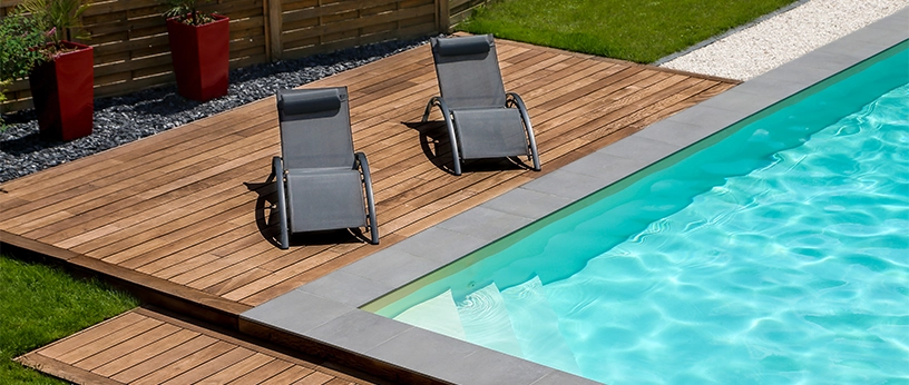 Poolterrasse mit Ipe-Holzdielen, zwei Sonnenliegen und modernem Randabschluss aus Beton, umgeben von Rasen und Gartenzaun.