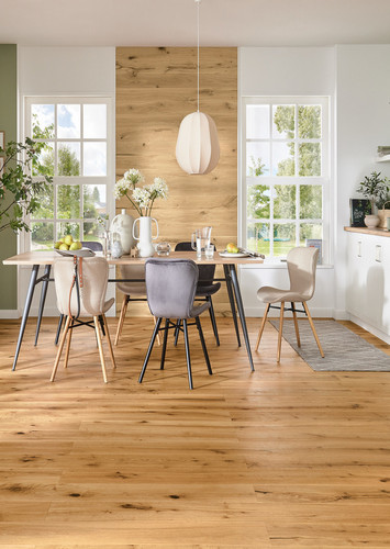 Landhaus-Wohnzimmer mit hellem Eichen-Holzboden, Ecksofa, Couchtisch, Regal und großen Fenstern mit Blick ins Grüne.