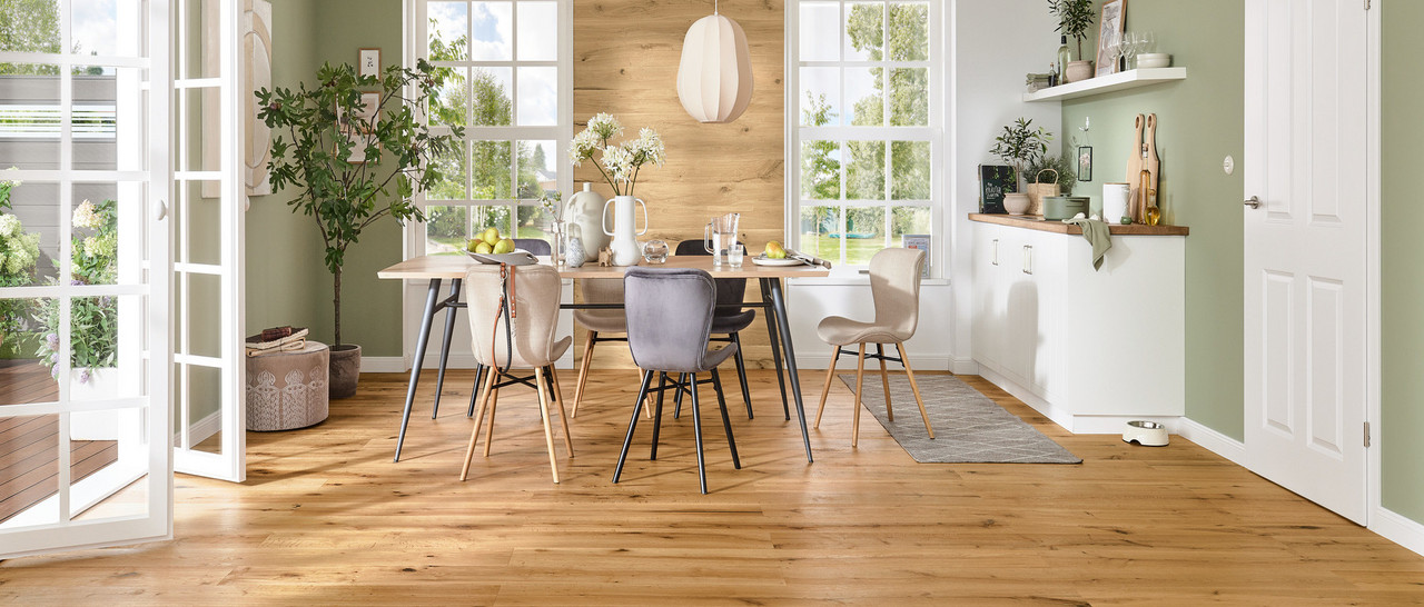 Landhaus-Wohnzimmer mit hellem Eichen-Holzboden, Ecksofa, Couchtisch, Regal und großen Fenstern mit Blick ins Grüne.