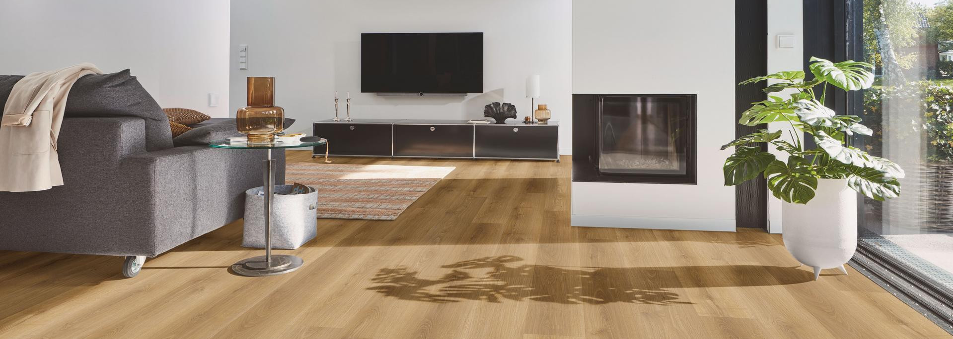 Wohnbereich mit modernem Designboden in warmer Eichenoptik, kombiniert mit Kamin, grauem Sofa und großen Fensterflächen.