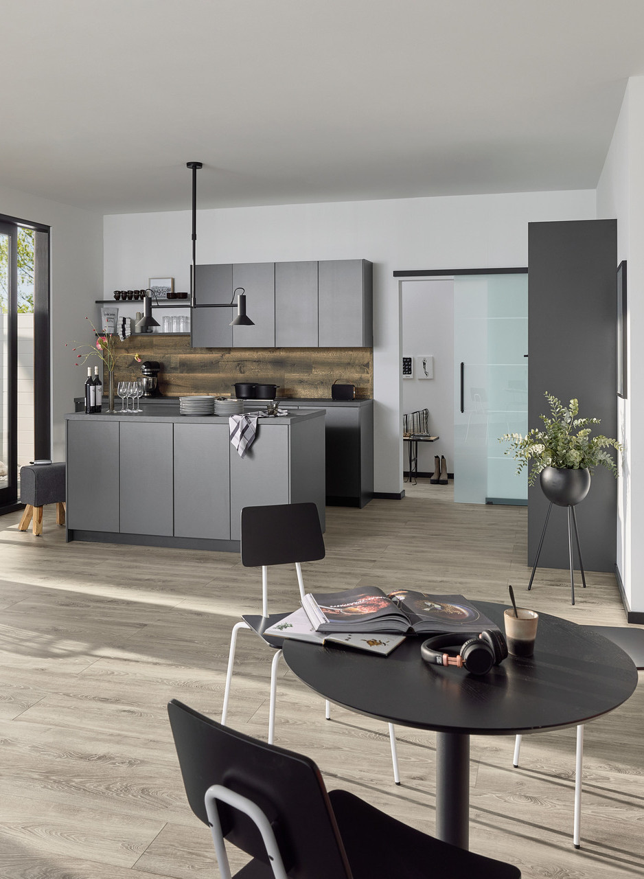Modernes Wohnküchen-Interieur mit grauem Holzboden, Kücheninsel, runder Esstischgruppe und großen Glasschiebetüren zur Terrasse.